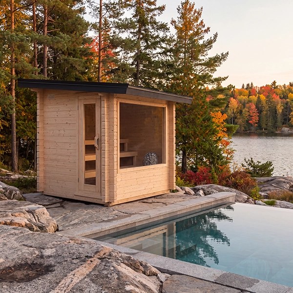 Avalon sauna with cedar walls, a large front window and black metal roof beside a lakeside pool.